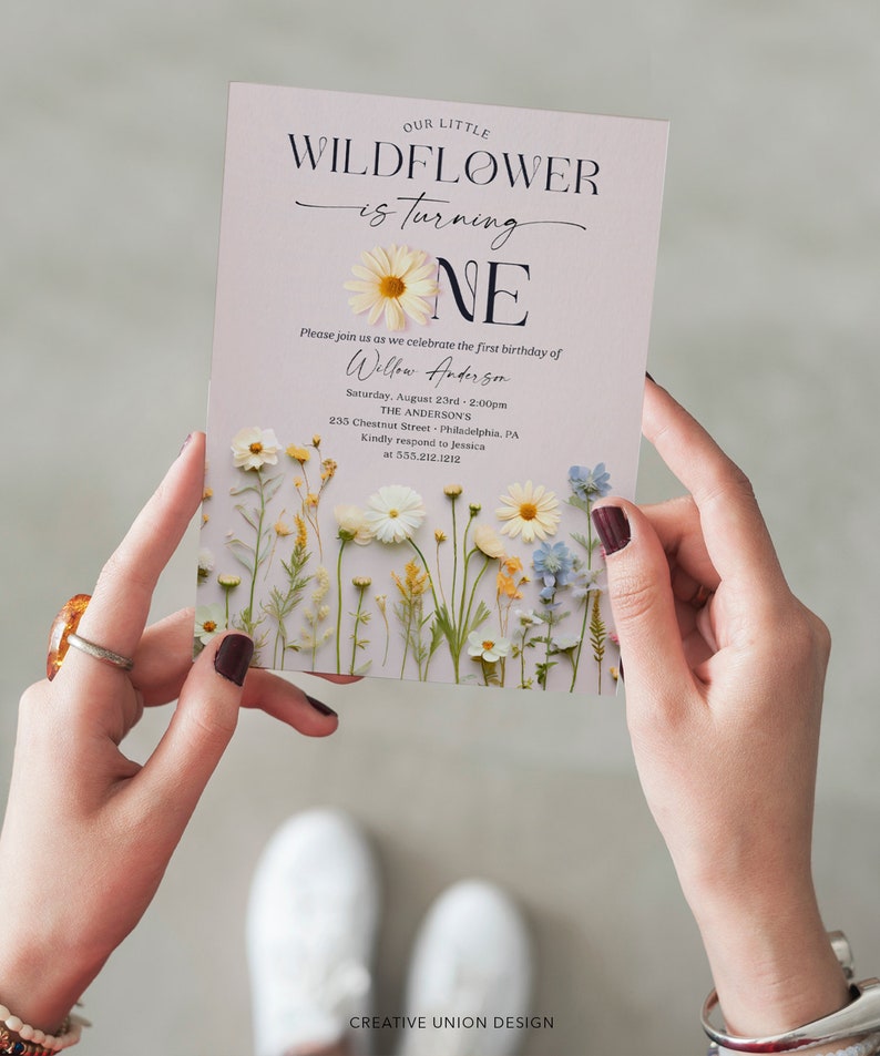 May include: A light purple invitation with a floral design featuring white daisies, yellow wildflowers, and blue flowers. The text reads "Our Little Wildflower is turning One" with a large white daisy and the number "One" in black text. The invitation includes the name "Willow Anderson", the date "Saturday, August 23rd - 2:00pm", the location "The Anderson's 235 Chestnut Street Philadelphia, PA", and the contact information "Kindly respond to Jessica at 535.212.1212". The bottom of the invitation reads "Creative Union Design".