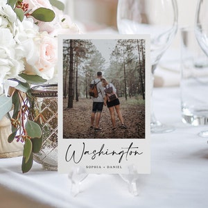 May include: A wedding table setting features a photo card with a couple in a forest, held by a clear acrylic stand. The card reads "Washington" in elegant script, with names below. Floral arrangements and glassware are in the background.