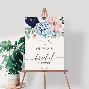 May include: A rectangular sign on a wooden easel featuring a floral arrangement of navy, light blue, and peach flowers at the top. The text reads: WELCOME TO OLIVIA'S bridal SHOWER 01.23.45. The background is off-white with dark gray lettering.