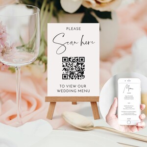 May include: A wedding menu sign on a small wooden easel. The sign is white with black text that reads "PLEASE Scan here" above a QR code and "TO VIEW OUR WEDDING MENU" below. A smartphone displays the menu.