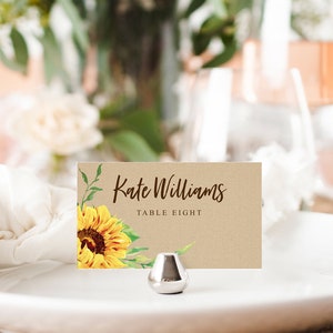 May include: A place card featuring a sunflower design and the text "Kate Williams, Table Eight." The card sits on a white plate, held by a silver metal stand. The background is blurred, with flowers and foliage.