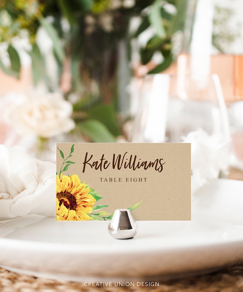 May include: A place card featuring a sunflower design and the text "Kate Williams, Table Eight." The card sits on a white plate, held by a silver metal stand. The background is blurred, with flowers and foliage.