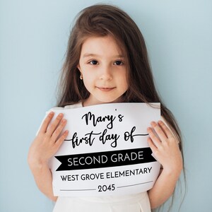May include: A white rectangular sign with black text that reads Mary's first day of SECOND GRADE WEST GROVE ELEMENTARY 2045. The sign features a mix of cursive and block lettering with a black banner graphic behind the words SECOND GRADE.