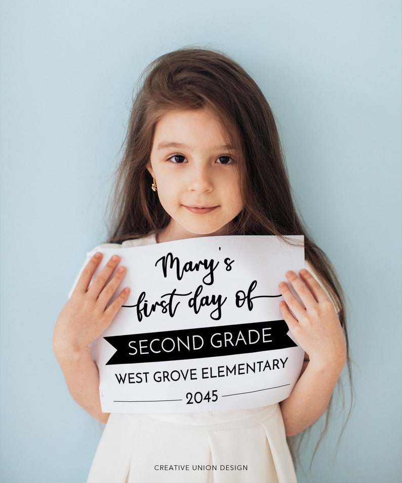 May include: A white rectangular sign with black text that reads Mary's first day of SECOND GRADE WEST GROVE ELEMENTARY 2045. The sign features a mix of cursive and block lettering with a black banner graphic behind the words SECOND GRADE.