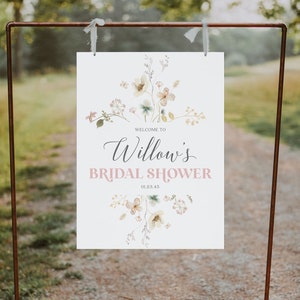 May include: A white sign with the text "Welcome to Willow's Bridal Shower" in elegant script and a soft pink font. Floral watercolor illustrations in pastel colors adorn the top and bottom. The sign is suspended from a copper-colored metal frame.