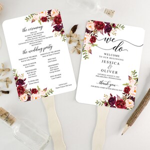 May include: Two white wedding fan programs with floral accents in burgundy, pink, and green. One fan reads "we do" with names and date. The other lists ceremony details and wedding party names. Wooden handles are attached.