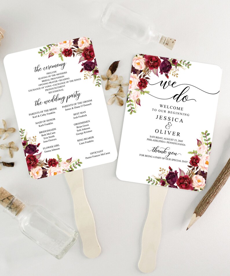 May include: Two white wedding fan programs with floral accents in burgundy, pink, and green. One fan reads "we do" with names and date. The other lists ceremony details and wedding party names. Wooden handles are attached.