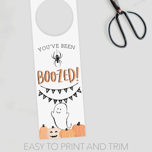 K&ouml;nnte beinhalten: Ein wei&szlig;er T&uuml;rh&auml;nger zum Thema Halloween mit einem kreisf&ouml;rmigen Ausschnitt oben. Das Design enth&auml;lt den Text "YOU'VE BEEN BOOZED!" mit einer Spinne, einem Banner mit der Aufschrift "HALLOWEEN", einem Geist und K&uuml;rbissen. Schwarze Scheren sind im Hintergrund.