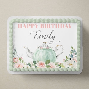 May include: A white cake topper with green frosting and a green and white teapot with pink flowers. The text "HAPPY BIRTHDAY Emily" is printed on the topper.
