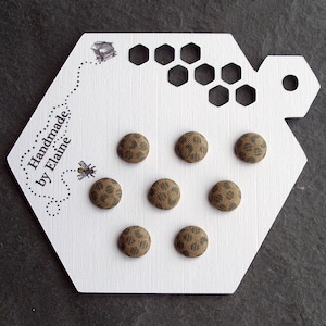 Op de afbeelding: Zeven bruine stoffen knopen met een zwart-wit geometrisch patroon. De knopen worden getoond op een witte zeshoekige kaart met de tekst "Handmade by Elaine".