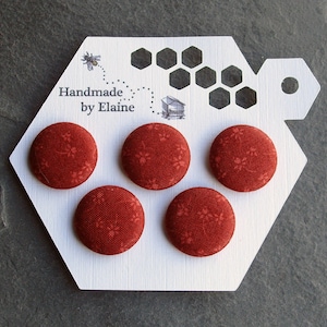 Peut inclure: Ensemble de quatre boutons en tissu rouge avec un motif floral blanc. Les boutons sont présentés sur une carte hexagonale blanche avec un motif d'abeille et de nid d'abeille. La carte comprend également le texte "Handmade by Elaine".