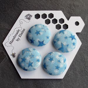 以下が含まれることがあります： 星柄の青と白の布ボタンが4つ。ボタンは「Handmade by Elaine」というテキストが書かれた白い六角形のカードに表示されています。