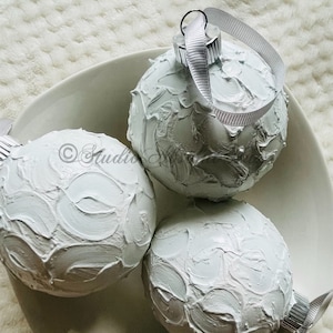 May include: Three white textured Christmas ornaments with silver-colored metal caps and white ribbon loops, resting in a white ceramic bowl. The ornaments have a thick, textured paint finish. The bowl sits on a white, textured surface.
