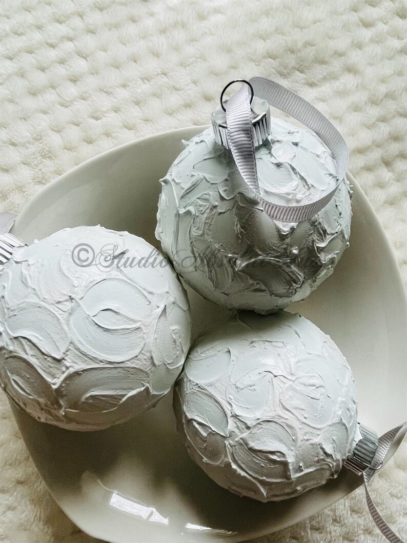 May include: Three white textured Christmas ornaments with silver-colored metal caps and white ribbon loops, resting in a white ceramic bowl. The ornaments have a thick, textured paint finish. The bowl sits on a white, textured surface.