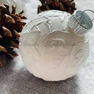 May include: A white Christmas ornament with a textured surface, a silver metal cap, and a metal loop for hanging. The ornament is next to a pine cone. The text "Studio Absolut Hues" is visible on the ornament.