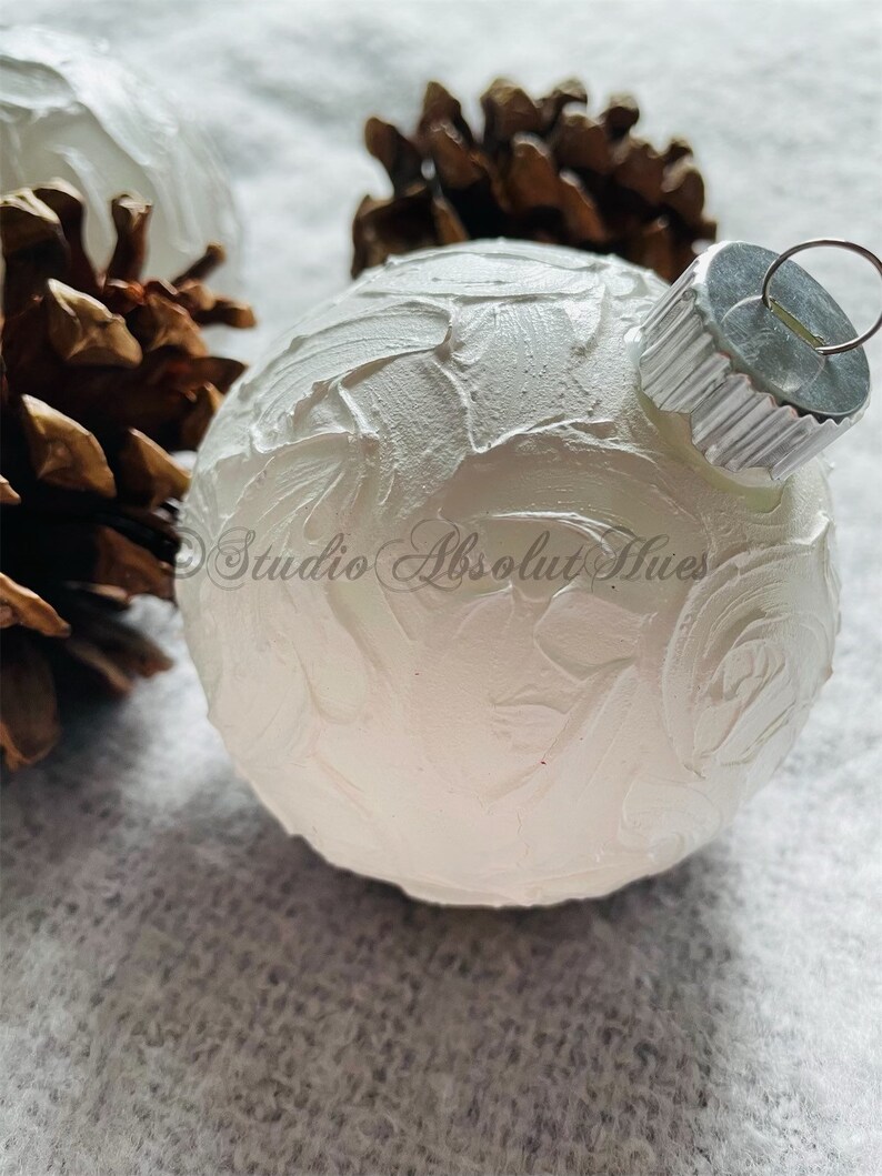 May include: A white Christmas ornament with a textured surface, a silver metal cap, and a metal loop for hanging. The ornament is next to a pine cone. The text "Studio Absolut Hues" is visible on the ornament.