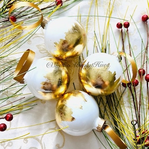 May include: Four white Christmas ornaments with gold leaf accents and gold ribbon hangers. The ornaments are arranged on a white patterned surface with sprigs of greenery and red berries.