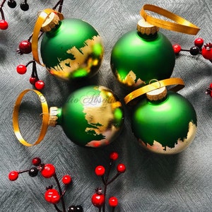 May include: Four green Christmas ornaments with gold accents and gold ribbon hangers. The ornaments are round and have a metallic finish. Red berries and branches are arranged around the ornaments.