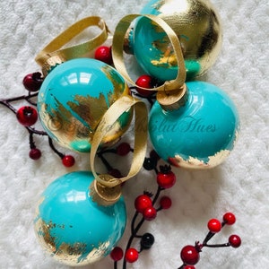 May include: Four turquoise Christmas ornaments with gold leaf accents and gold ribbon hangers. The ornaments are arranged with red berries on a white, textured surface, creating a festive holiday display.