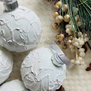 May include: White textured Christmas ornaments with silver ribbon hangers. The ornaments have a textured, painted surface. The image also includes pine cones, berries, and evergreen branches, creating a festive holiday theme.