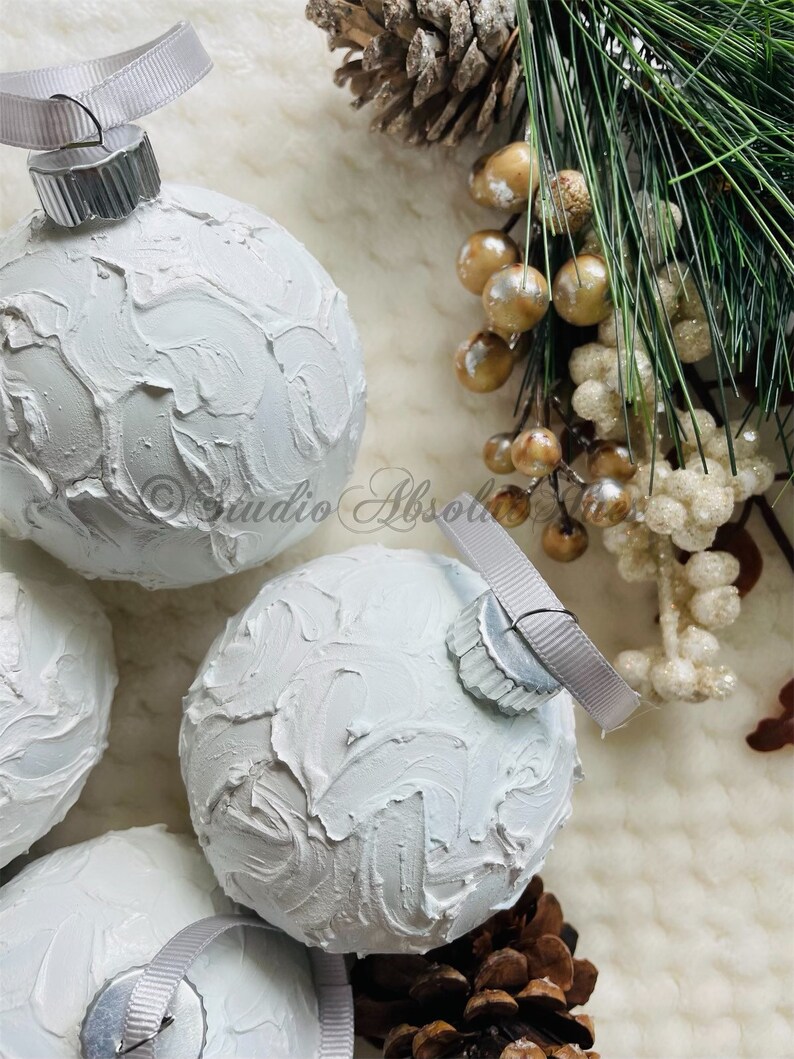 May include: White textured Christmas ornaments with silver ribbon hangers. The ornaments have a textured, painted surface. The image also includes pine cones, berries, and evergreen branches, creating a festive holiday theme.