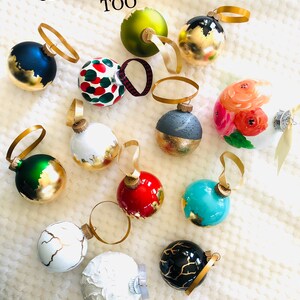 May include: A collection of decorative Christmas ornaments in various colors, including blue, green, red, and white, with gold accents. Some ornaments feature painted designs, and all have gold ribbon hangers. The text "CHECK THESE OUT TOO" is visible.