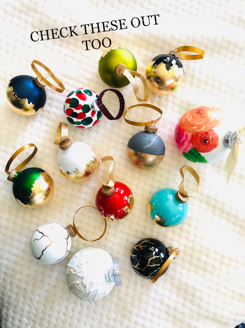 May include: A collection of decorative Christmas ornaments in various colors, including blue, green, red, and white, with gold accents. Some ornaments feature painted designs, and all have gold ribbon hangers. The text "CHECK THESE OUT TOO" is visible.