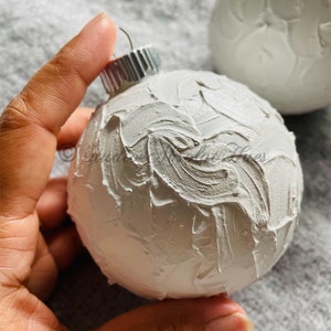 May include: White Christmas ornament with a textured, layered paint effect. The ornament has a silver metal cap and a loop for hanging. Another ornament is visible in the background. The ornaments are on a gray surface.