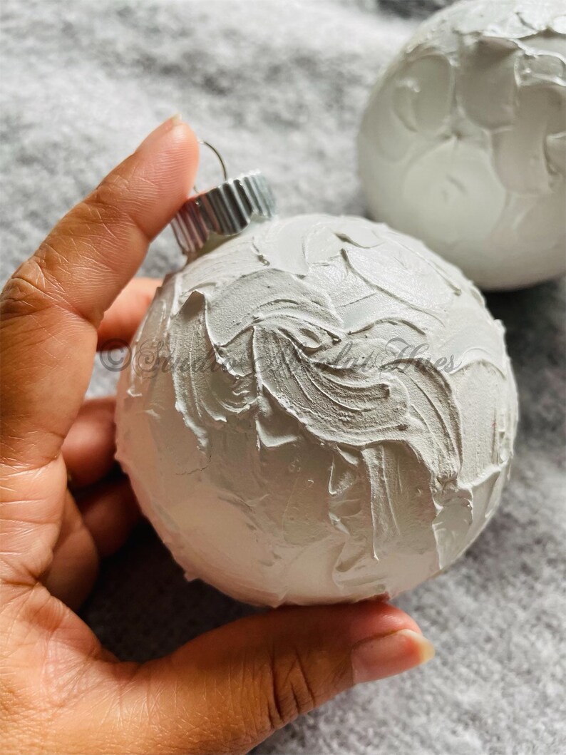 May include: White Christmas ornament with a textured, layered paint effect. The ornament has a silver metal cap and a loop for hanging. Another ornament is visible in the background. The ornaments are on a gray surface.