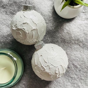 May include: Two white Christmas ornaments with textured surfaces and silver metal caps. A small white ceramic pot with a green plant and a green glass candle holder are also visible.