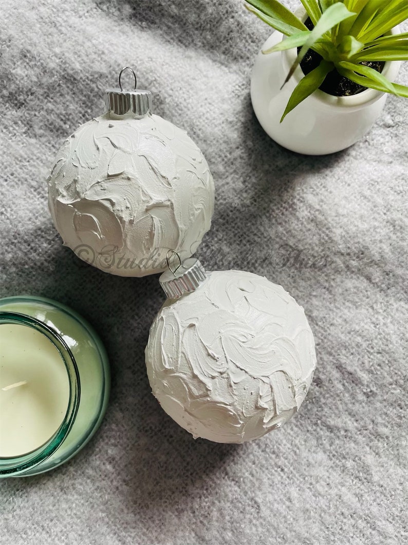 May include: Two white Christmas ornaments with textured surfaces and silver metal caps. A small white ceramic pot with a green plant and a green glass candle holder are also visible.