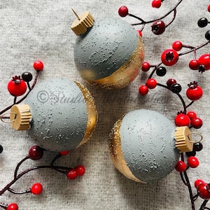 May include: Three gray and gold Christmas ornaments with textured surfaces. Each ornament has a gold bottom half and a wooden cap. Red berries and branches are arranged around the ornaments.