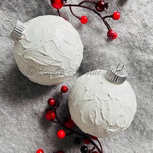 May include: Two white Christmas ornaments with textured surfaces and silver metal caps are displayed. Red and black berries on brown stems are arranged around the ornaments, set against a gray background. The ornaments are a festive holiday decoration.