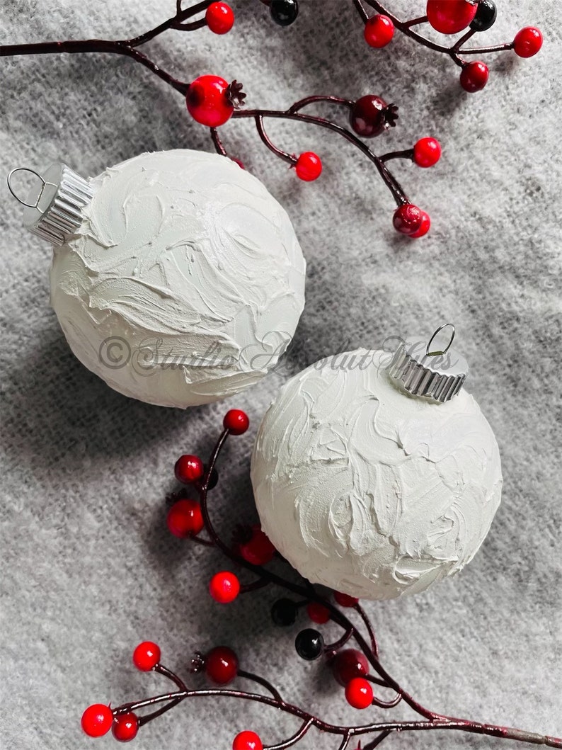 May include: Two white Christmas ornaments with textured surfaces and silver metal caps are displayed. Red and black berries on brown stems are arranged around the ornaments, set against a gray background. The ornaments are a festive holiday decoration.