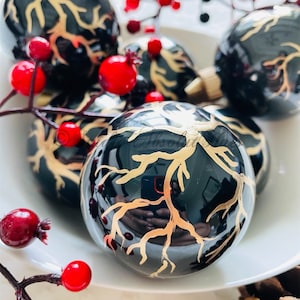 May include: Black Christmas ornaments with gold branch designs are arranged in a white bowl, accented by red berries. The glossy ornaments reflect light, creating a festive holiday display. A pine cone is visible in the background.
