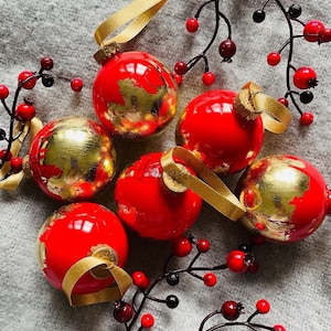 May include: Red Christmas ornaments with gold leaf accents and gold ribbon hangers are arranged with sprigs of red and black berries. The ornaments are round and glossy, creating a festive holiday display.