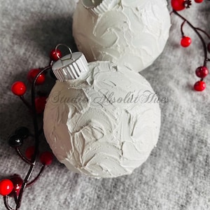 May include: Two white textured Christmas ornaments with silver caps, resting on a gray surface. Red berries and dark stems are arranged around the ornaments, adding a festive touch. The ornaments have a unique, handcrafted appearance.