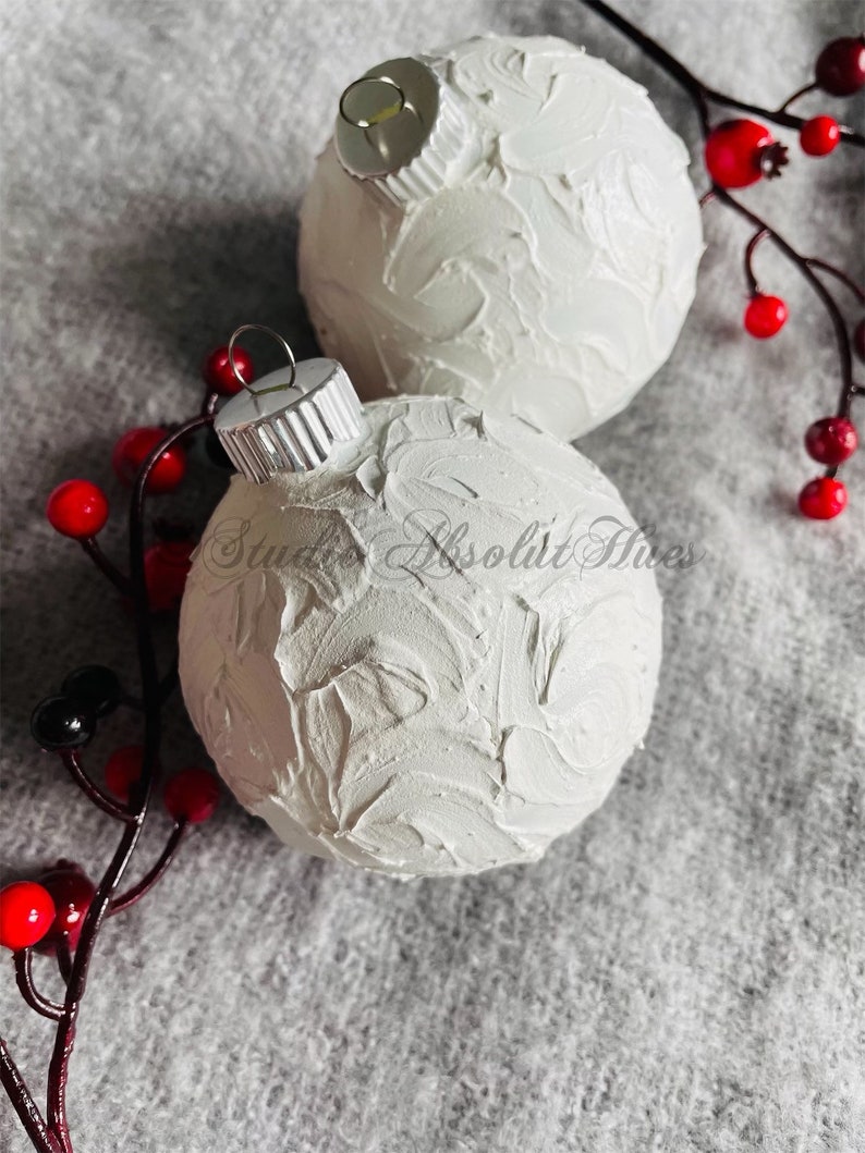 May include: Two white textured Christmas ornaments with silver caps, resting on a gray surface. Red berries and dark stems are arranged around the ornaments, adding a festive touch. The ornaments have a unique, handcrafted appearance.