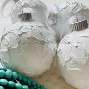 May include: Close-up of four white textured Christmas ornaments with silver caps and white ribbon hangers. A strand of turquoise beads is in the foreground. The ornaments have a textured, painted surface.