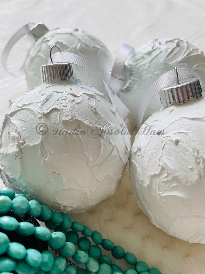 May include: Close-up of four white textured Christmas ornaments with silver caps and white ribbon hangers. A strand of turquoise beads is in the foreground. The ornaments have a textured, painted surface.