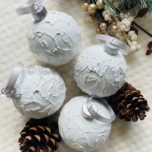 May include: Four white textured Christmas ornaments with silver ribbon hangers. The ornaments are arranged with pine cones and greenery, creating a festive holiday display. The ornaments have a textured, painted surface.