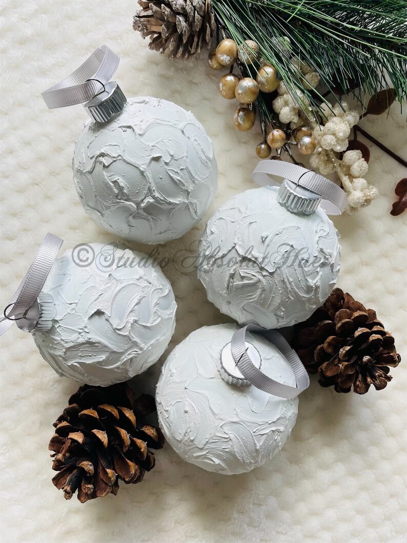 May include: Four white textured Christmas ornaments with silver ribbon hangers. The ornaments are arranged with pine cones and greenery, creating a festive holiday display. The ornaments have a textured, painted surface.