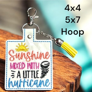Puede incluir: Un llavero blanco con un diseño bordado de colores que dice "Sunshine mixed with a little hurricane". El llavero tiene un borla amarilla.