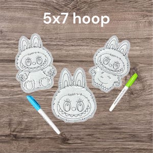Könnte beinhalten: Drei weiß gestickte Monster-Hasen-Designs mit schwarzen Umrissen, jedes mit einem einzigartigen Ausdruck. Der Text "5x7 hoop" befindet sich über den Designs. Zwei Marker, einer blau und einer grün, liegen auf der Holzoberfläche.