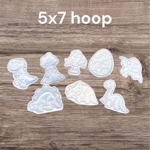 May include: A collection of light blue dinosaur-themed appliqués, including a T-Rex, a Triceratops, and a Brontosaurus, along with a volcano, trees, and an egg. The text "5x7 hoop" is at the top. These are likely for crafting or embroidery projects.