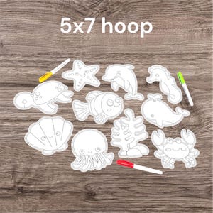 May include: A collection of white, outline-style sea creature cutouts, including a turtle, starfish, dolphin, seahorse, fish, whale, crab, jellyfish, seashell, and coral. The text "5x7 hoop" is at the top. Three markers are also present.