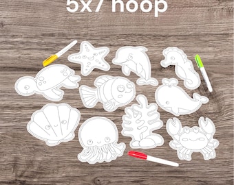 May include: A collection of white fabric cutouts featuring black line art of sea creatures including a starfish, dolphin, seahorse, turtle, fish, whale, jellyfish, crab, and coral, with the text 5x7 hoop and three markers in yellow, green, and red.