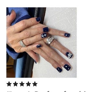 May include: Manicured fingernails painted in a glossy dark navy blue polish. The nails feature white accent designs, including a nautical anchor symbol on one finger and a star pattern on another. Two silver-toned rings with clear stones are worn.