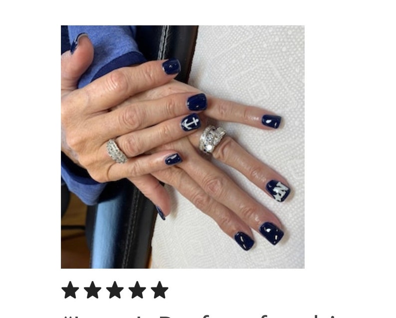 May include: Manicured fingernails painted in a glossy dark navy blue polish. The nails feature white accent designs, including a nautical anchor symbol on one finger and a star pattern on another. Two silver-toned rings with clear stones are worn.