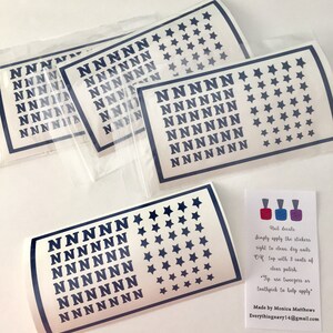 May include: Four rectangular sheets of dark blue nail decals featuring a repeating pattern of the letter N and five-pointed stars, enclosed in clear plastic sleeves. A small white instruction card with three nail polish bottle icons is included.